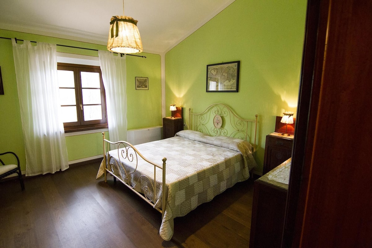 Un dormitorio con cama, dos mesitas de noche, una ventana con cortinas y una lámpara colgante.