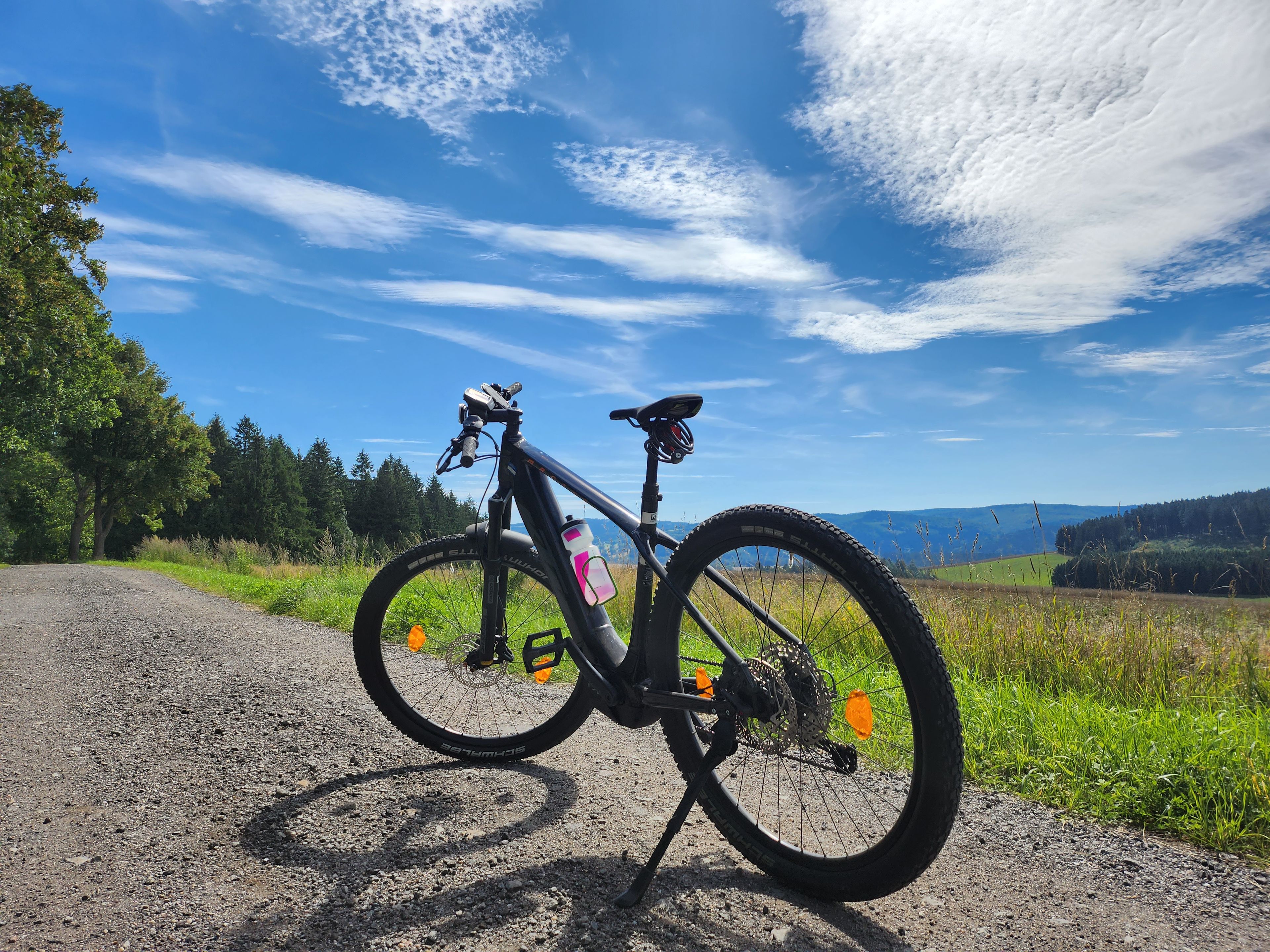 Una bicicleta de montaña estacionada en un sendero de grava, con una vista panorámica de un bosque y colinas al fondo.