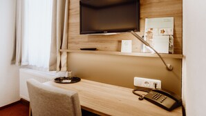 A hotel room with a wooden desk, a flat-screen TV mounted on the wall, a telephone, and a small shelf with a coffee maker and some items. - Hotel Frauensteiner Hof (Frauenstein)