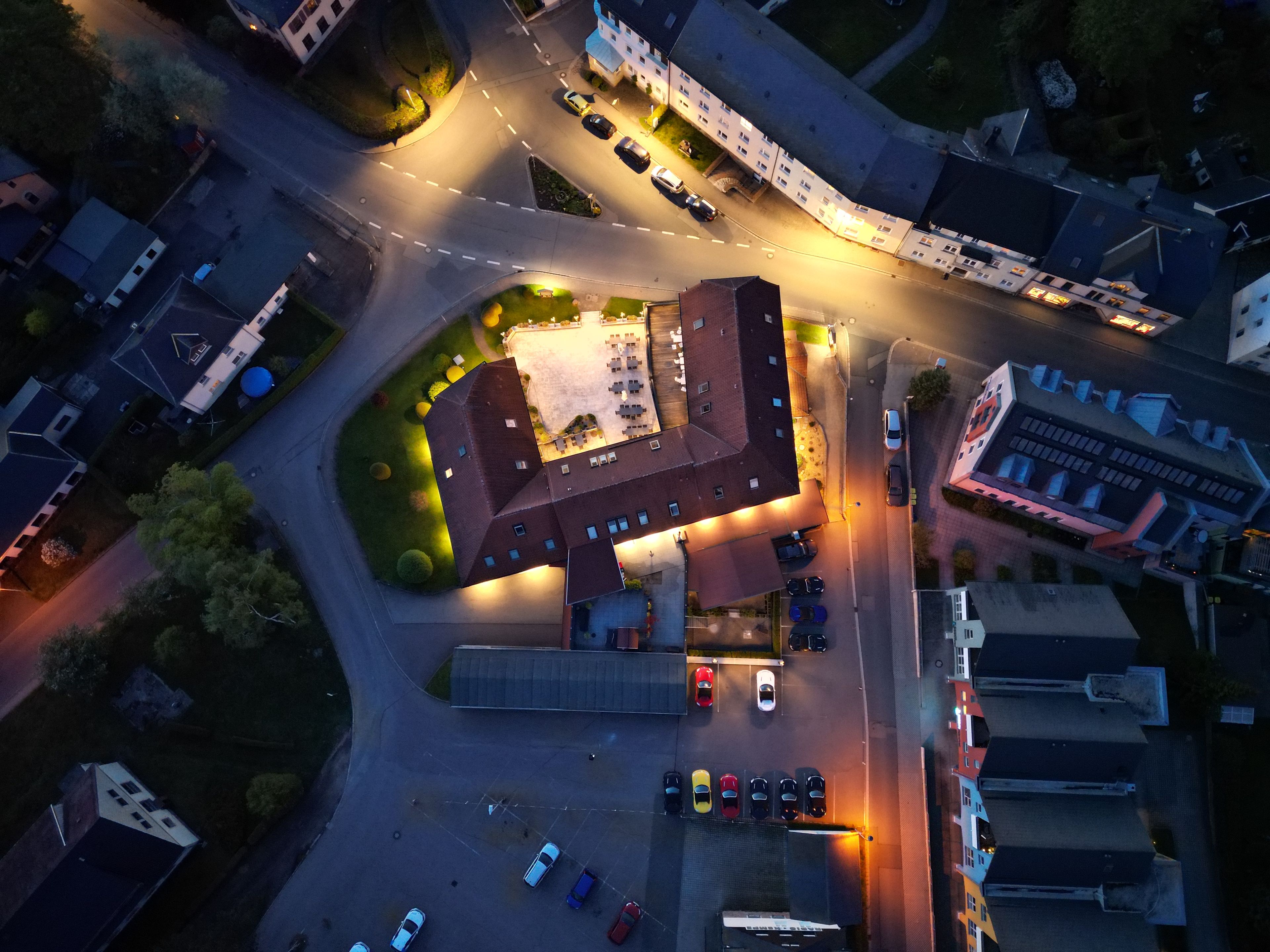 Eine Luftaufnahme eines Parkplatzes mit mehreren Autos, einem Gebäude mit markanter Architektur und einem gut beleuchteten Bereich mit Außenbestuhlung.