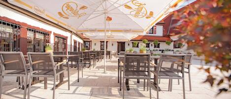 An outdoor seating area with tables and chairs under large umbrellas, a building with a red-tiled roof, and a clear sky.