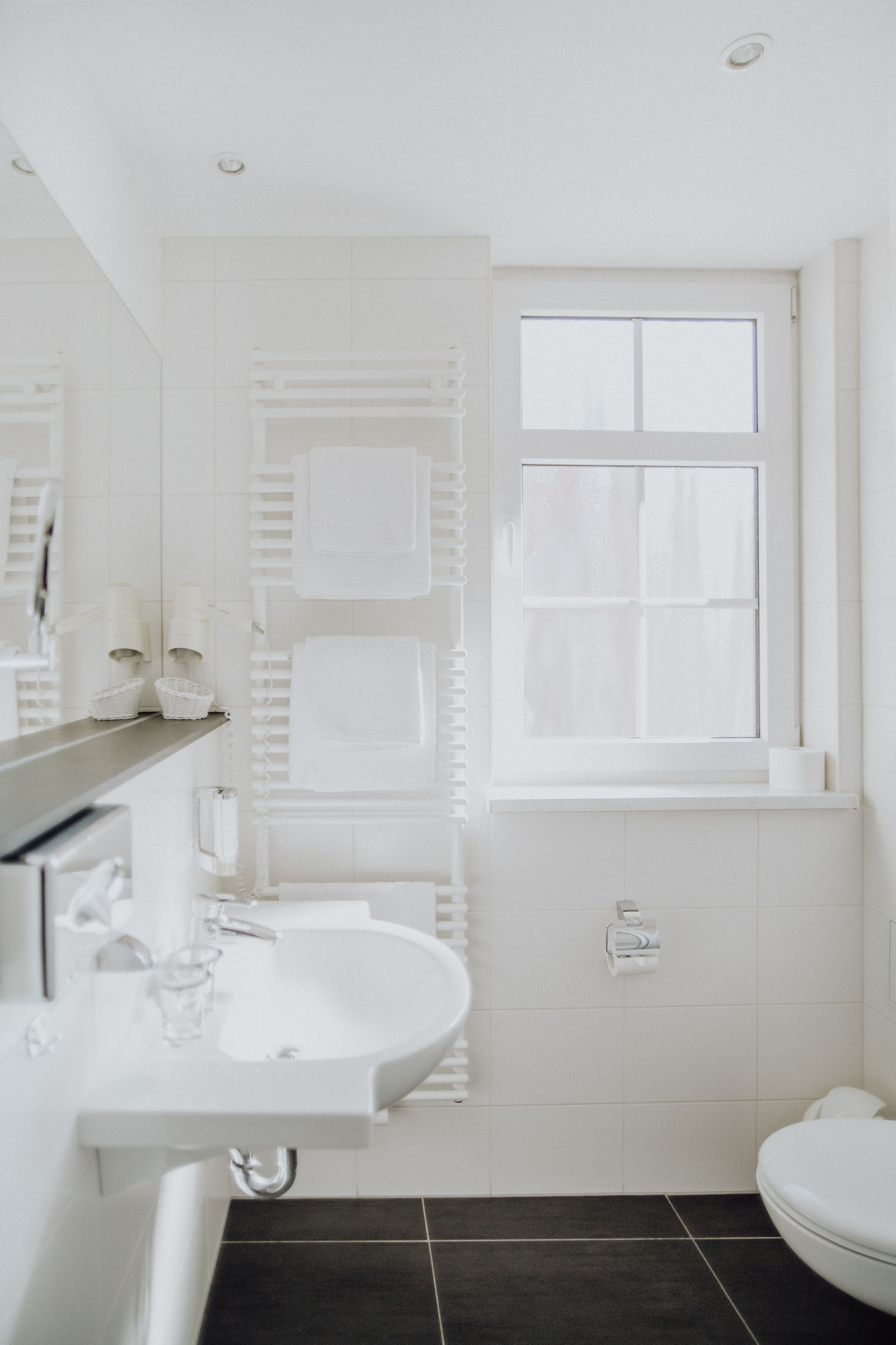 Ein modernes Badezimmer mit Waschbecken, Spiegel und Toilette in Weiß. Über der Toilette befindet sich ein Fenster und an der Wand ein Handtuchhalter.
