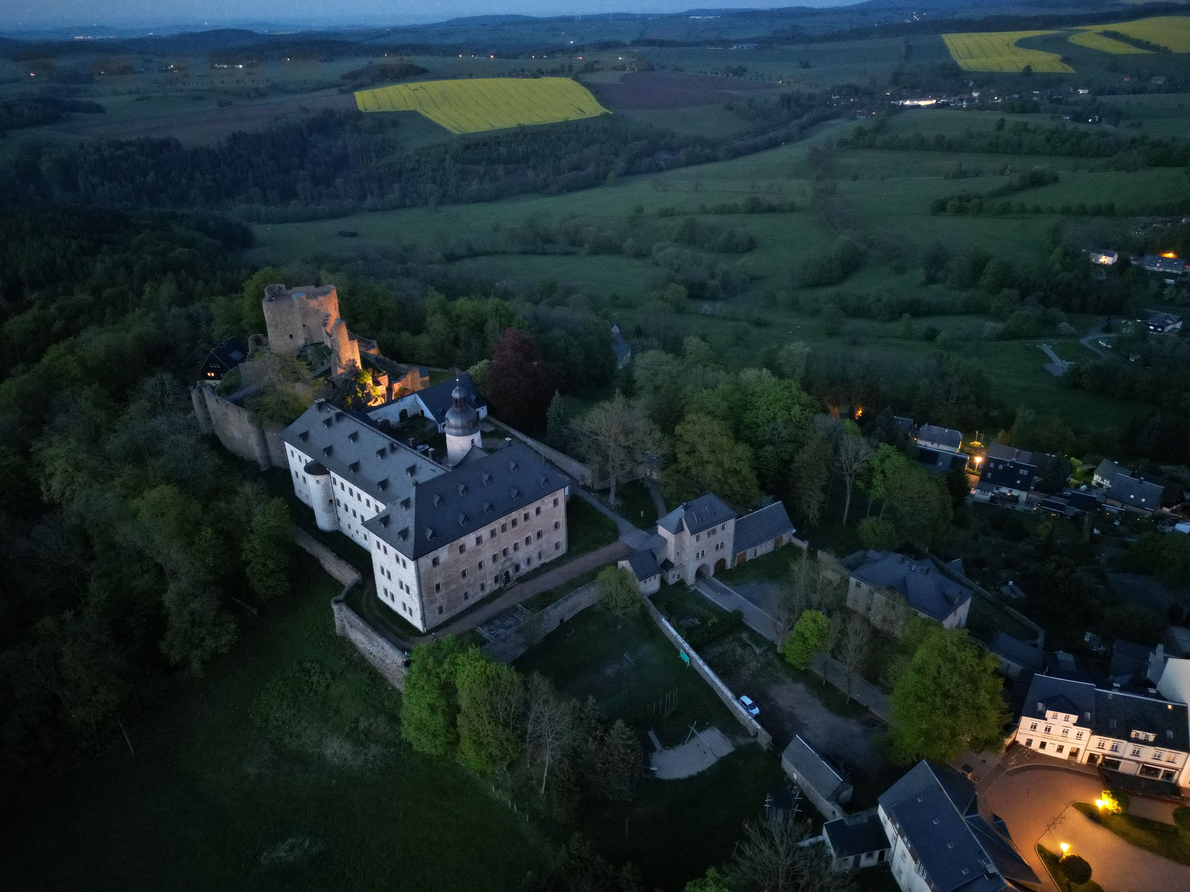 An aerial view of a castle complex with multiple buildings, surrounded by fields and trees.