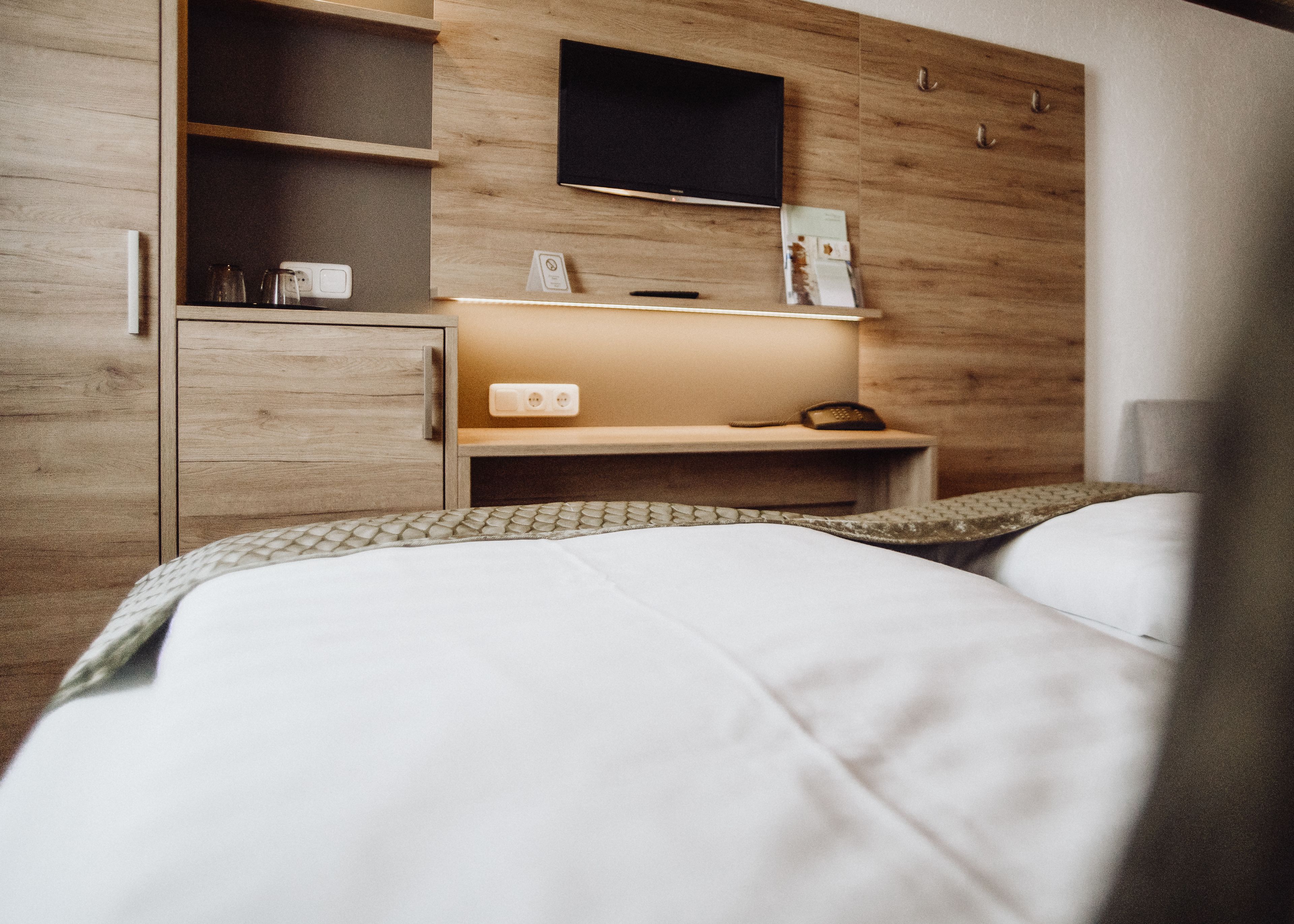 A modern hotel room with a wooden wardrobe, a bedside table with a lamp, and a flat-screen TV mounted on the wall.