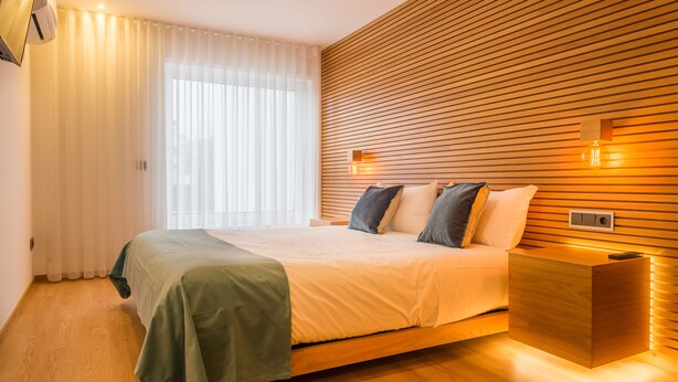 A modern bedroom with a wooden slat wall, a bed with a green bedspread, and a bedside table with a lamp. - Almada Domus (Porto)