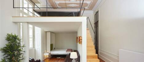 A spacious loft apartment with a high ceiling, a staircase, a bed, a desk, and a seating area.