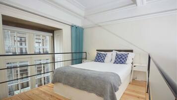 Un dormitorio con cama, mesita de noche, ventana con vistas a edificios y barandilla de balcón.