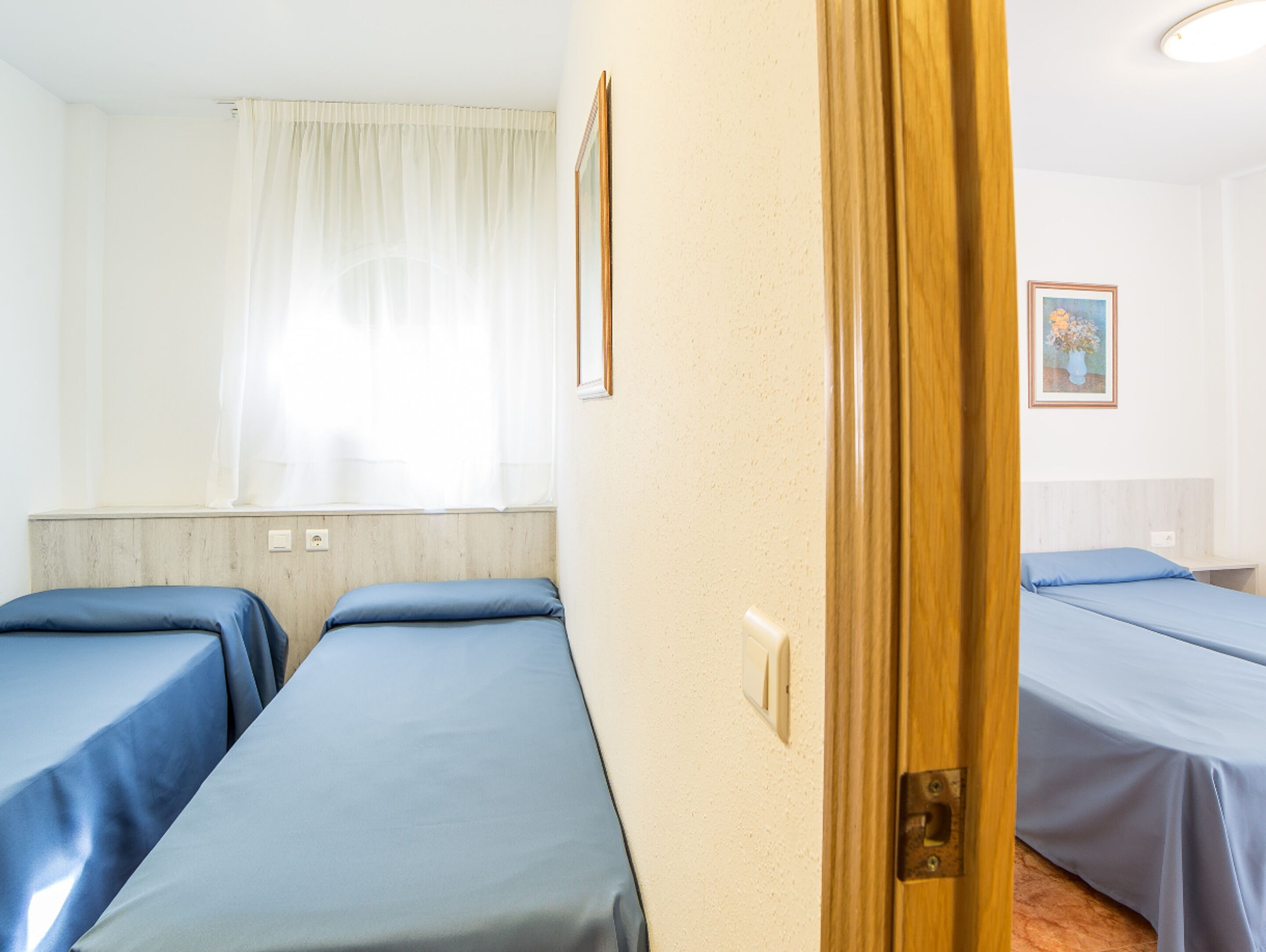 A hotel room with two single beds, a wooden door, a light switch, and a framed picture on the wall.