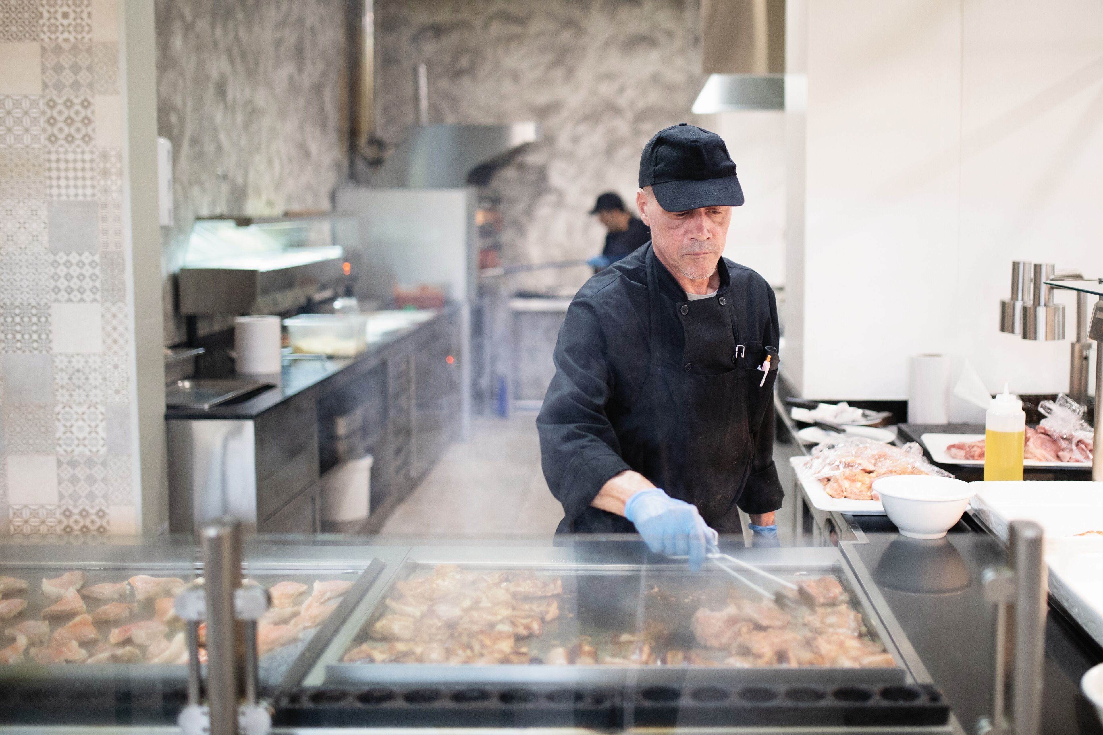 Une personne est en train de faire griller de la viande dans une cuisine dont le mur derrière le plan de travail est recouvert de carreaux et qui est équipée d’appareils électroménagers en acier inoxydable.