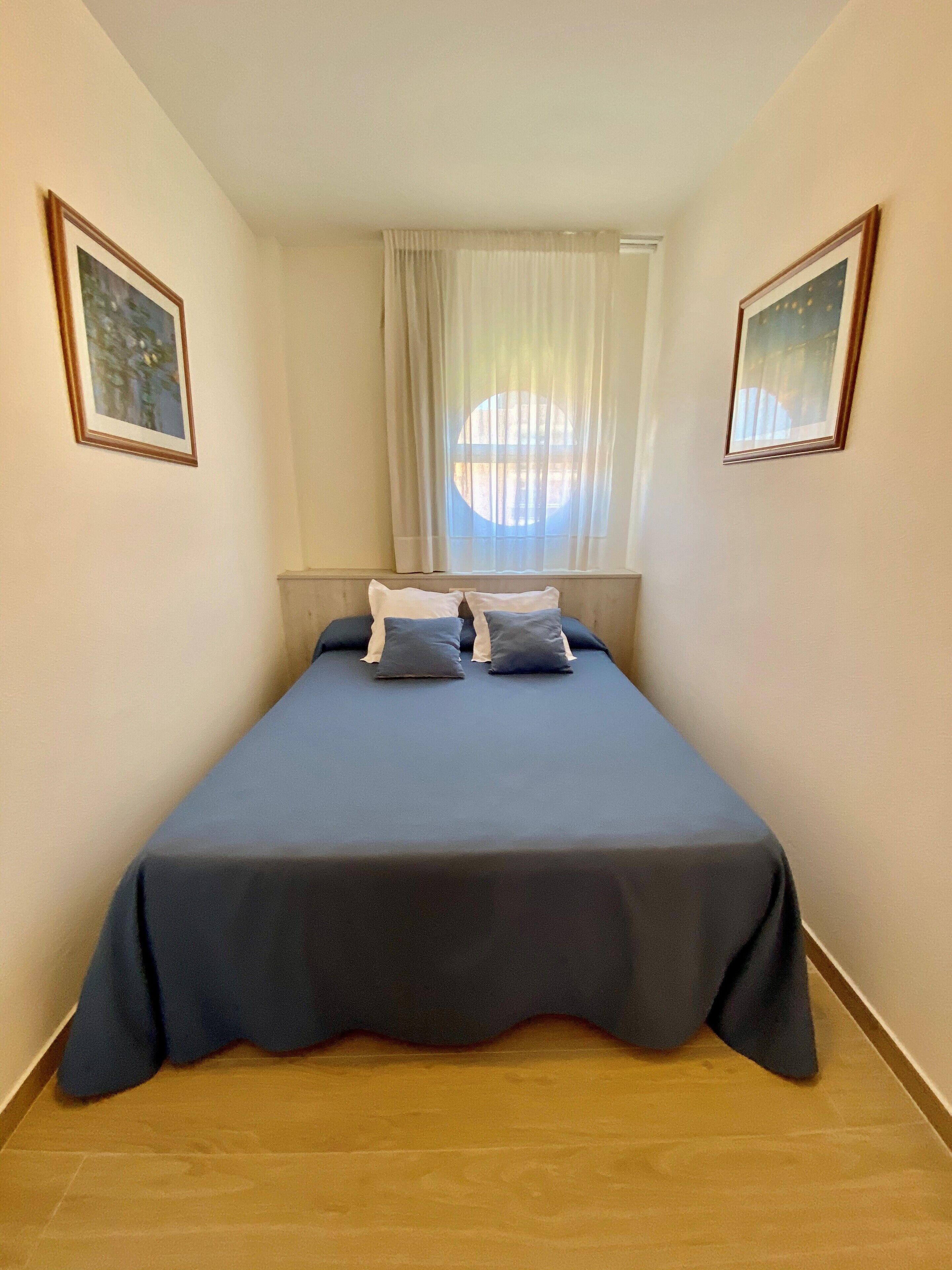 Un petit lit bien fait, avec une couverture bleue et deux oreillers, une fenêtre ornée de rideaux légers et deux tableaux encadrés accrochés aux murs.