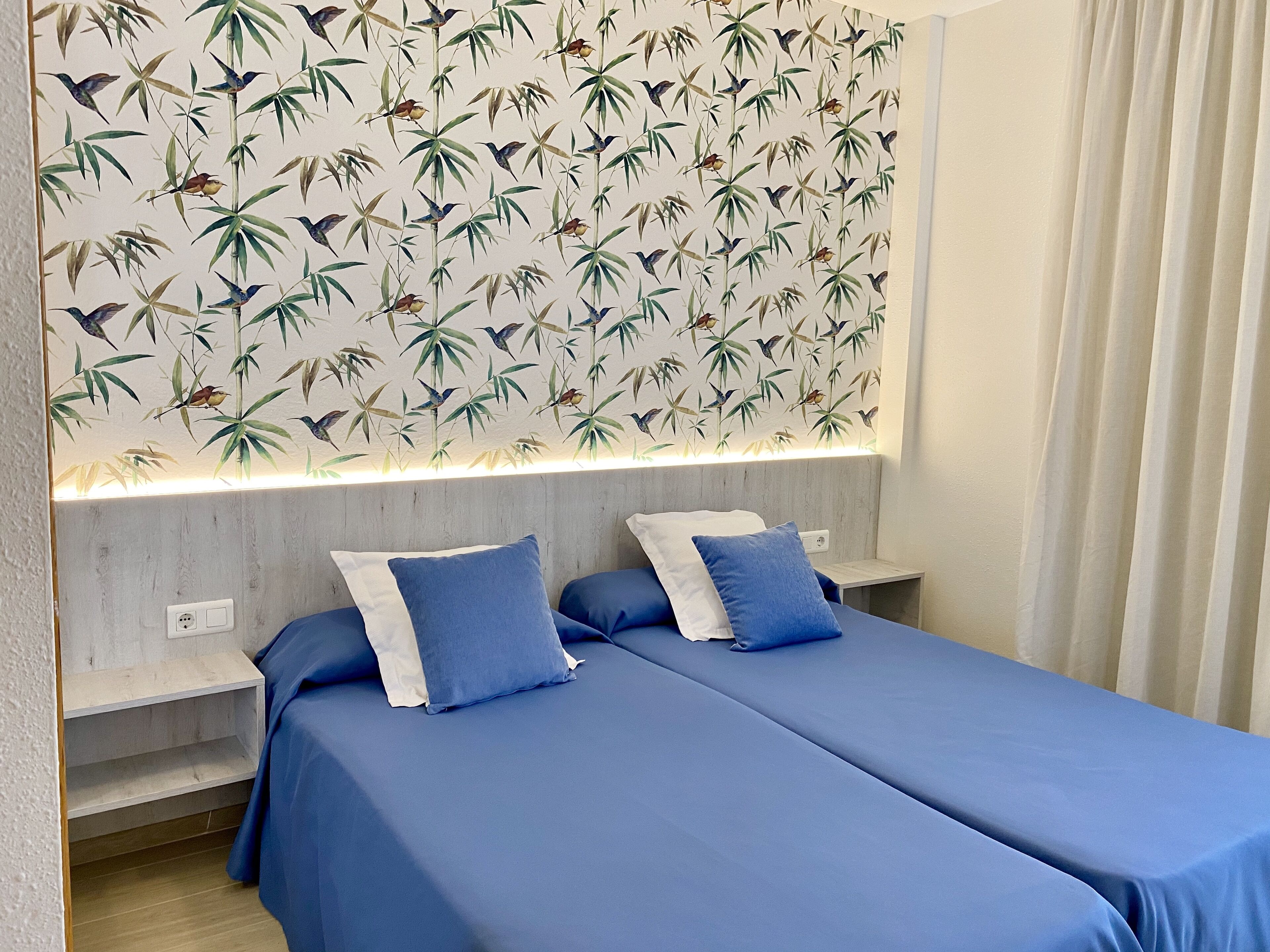 A room with two beds, each with blue bedding and white pillows. The wall above the beds features a bamboo leaf and bird pattern.