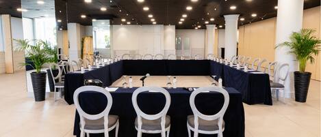 Un salón de conferencias con una larga mesa, sillas blancas y plantas en macetas.