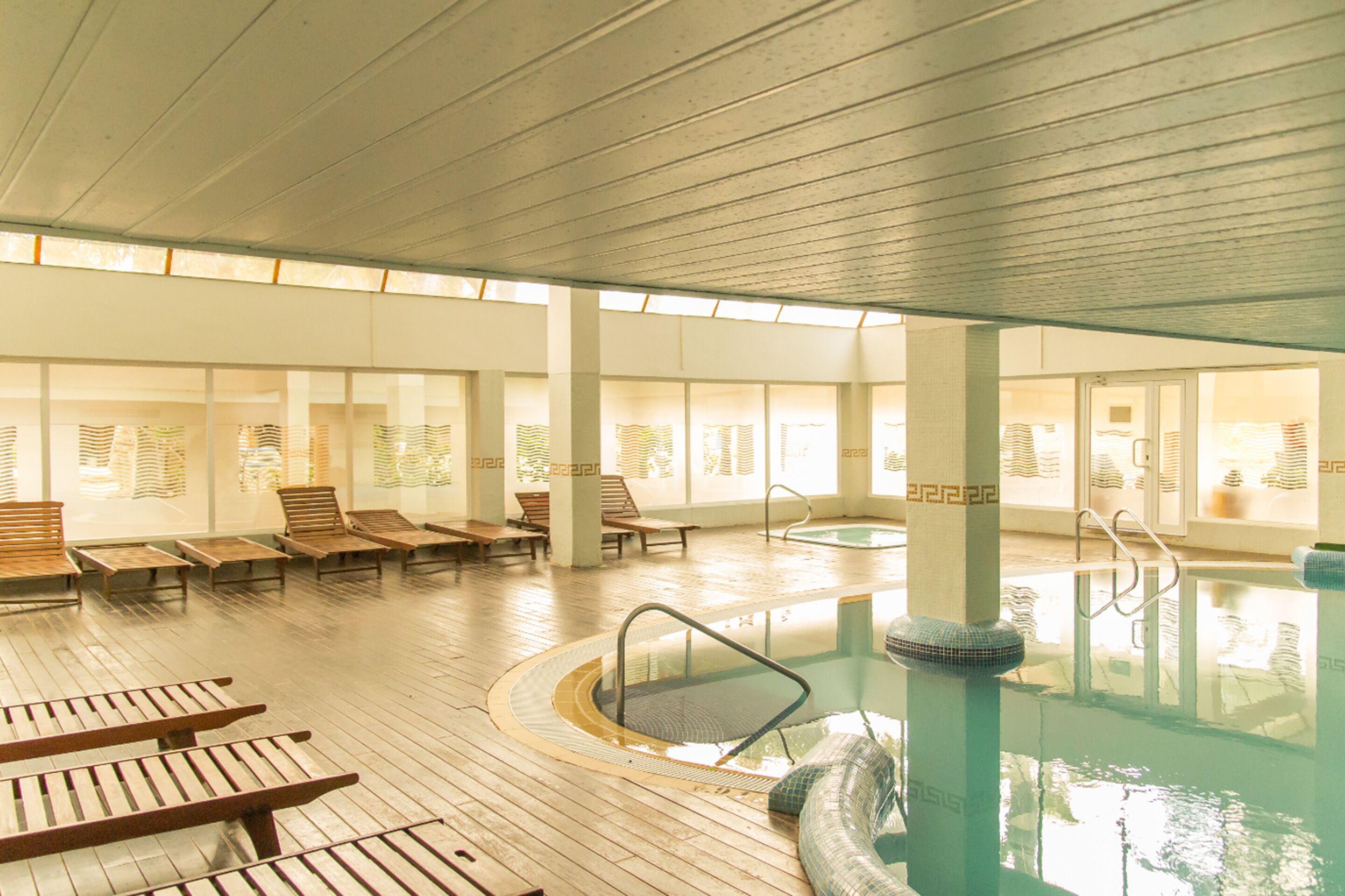 A large indoor swimming pool with wooden loungers and a sunlit room with large windows.