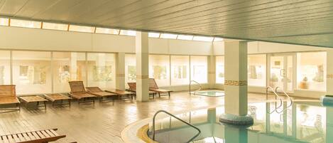 A large indoor swimming pool with wooden loungers and a sunlit room with large windows.