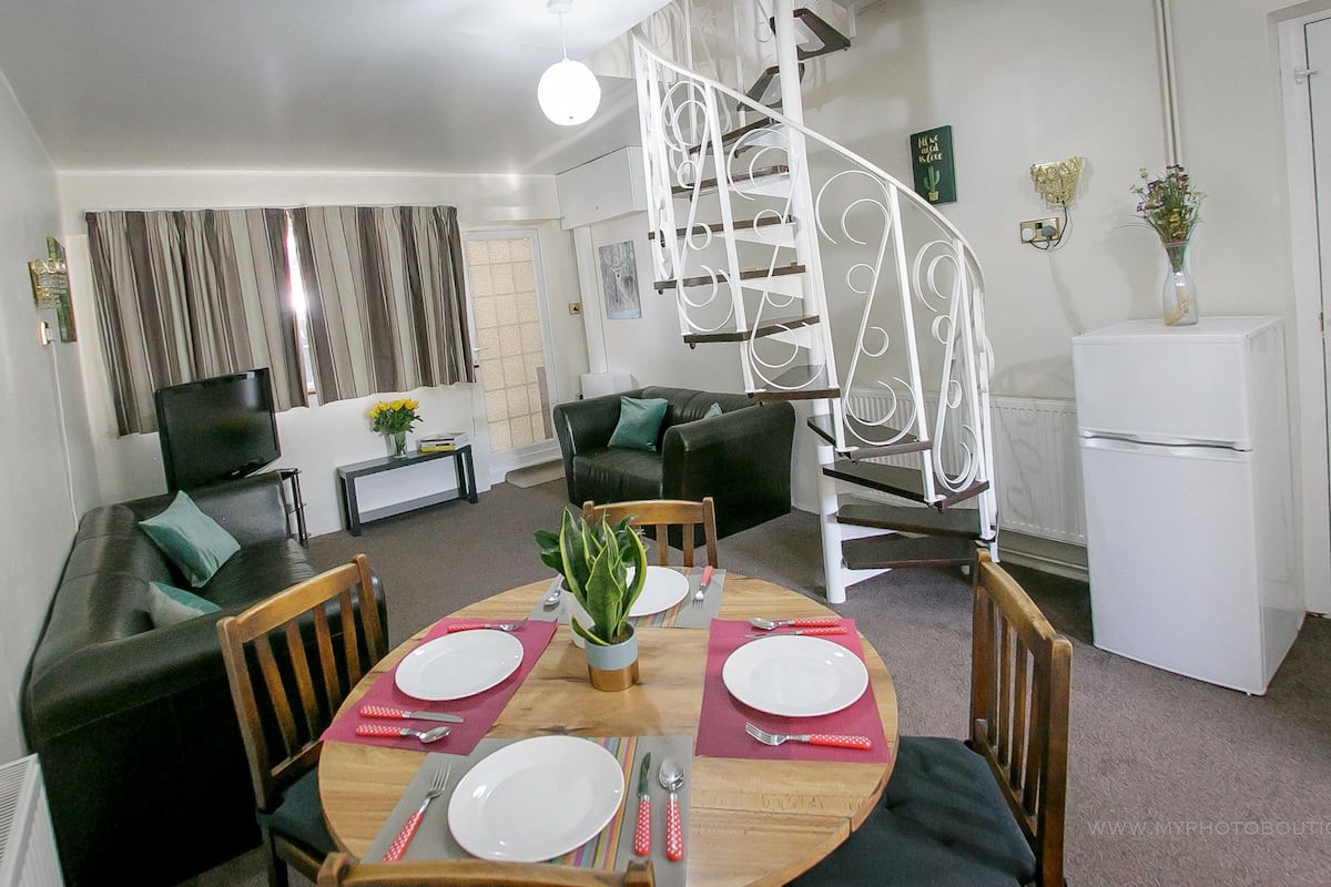 Un coin repas avec une table ronde dressée pour quatre personnes, entourée de chaises en bois. Un escalier en colimaçon est visible au fond.