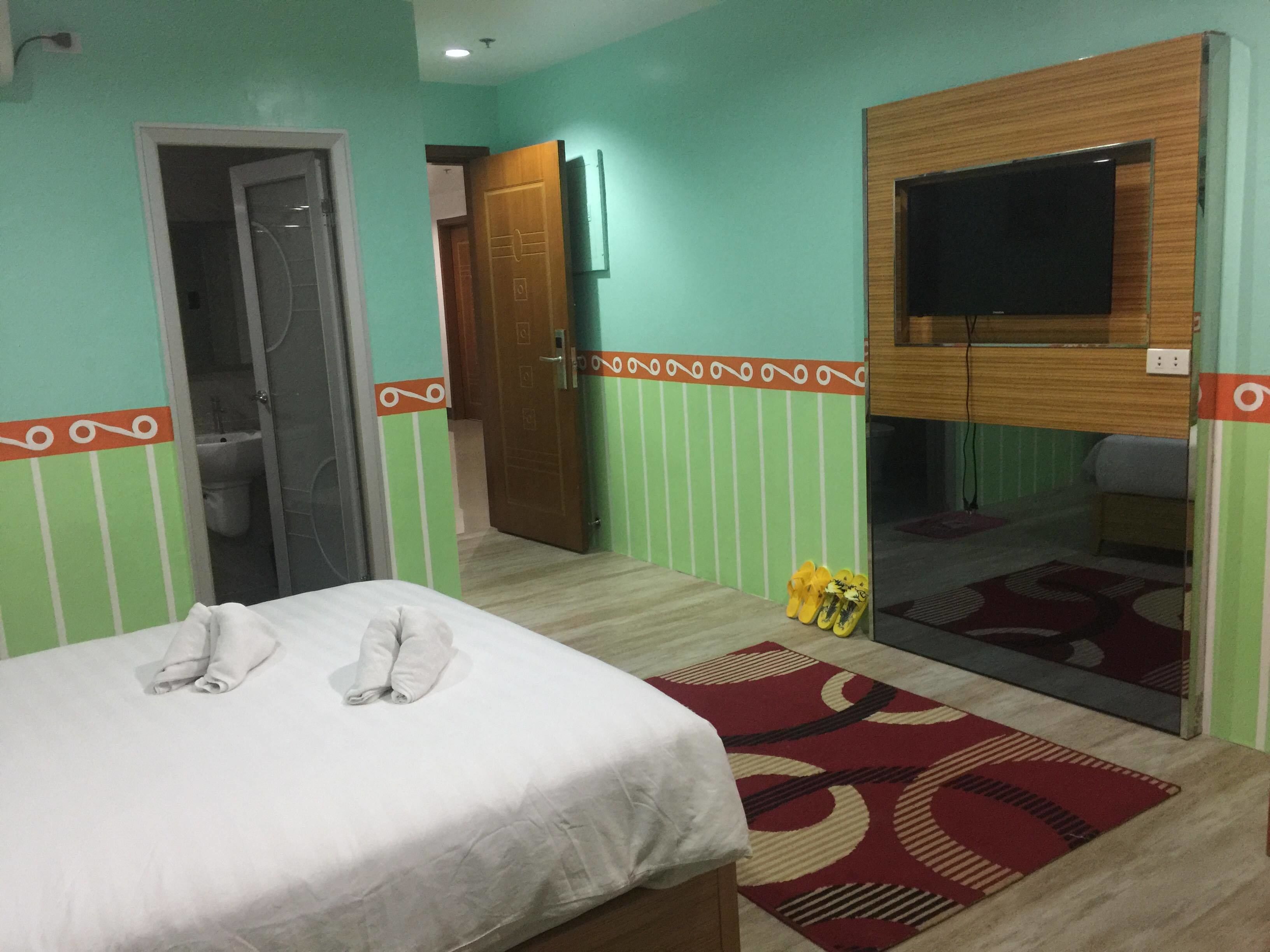 Een hotelkamer met een bed, een aan de muur bevestigde televisie, een badkamer zichtbaar door een open deur, en een paar slippers op de vloer.
