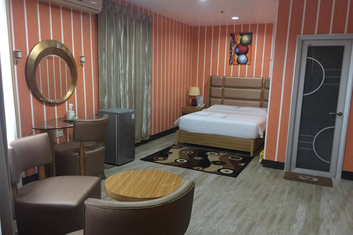 Een hotelkamer met een bed, een bureau, een stoel, een tafeltje, een koelkast en een deur met een raam.
