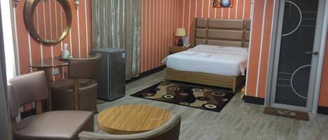 A hotel room with a bed, a desk, a chair, a small table, a refrigerator, and a door with a window.