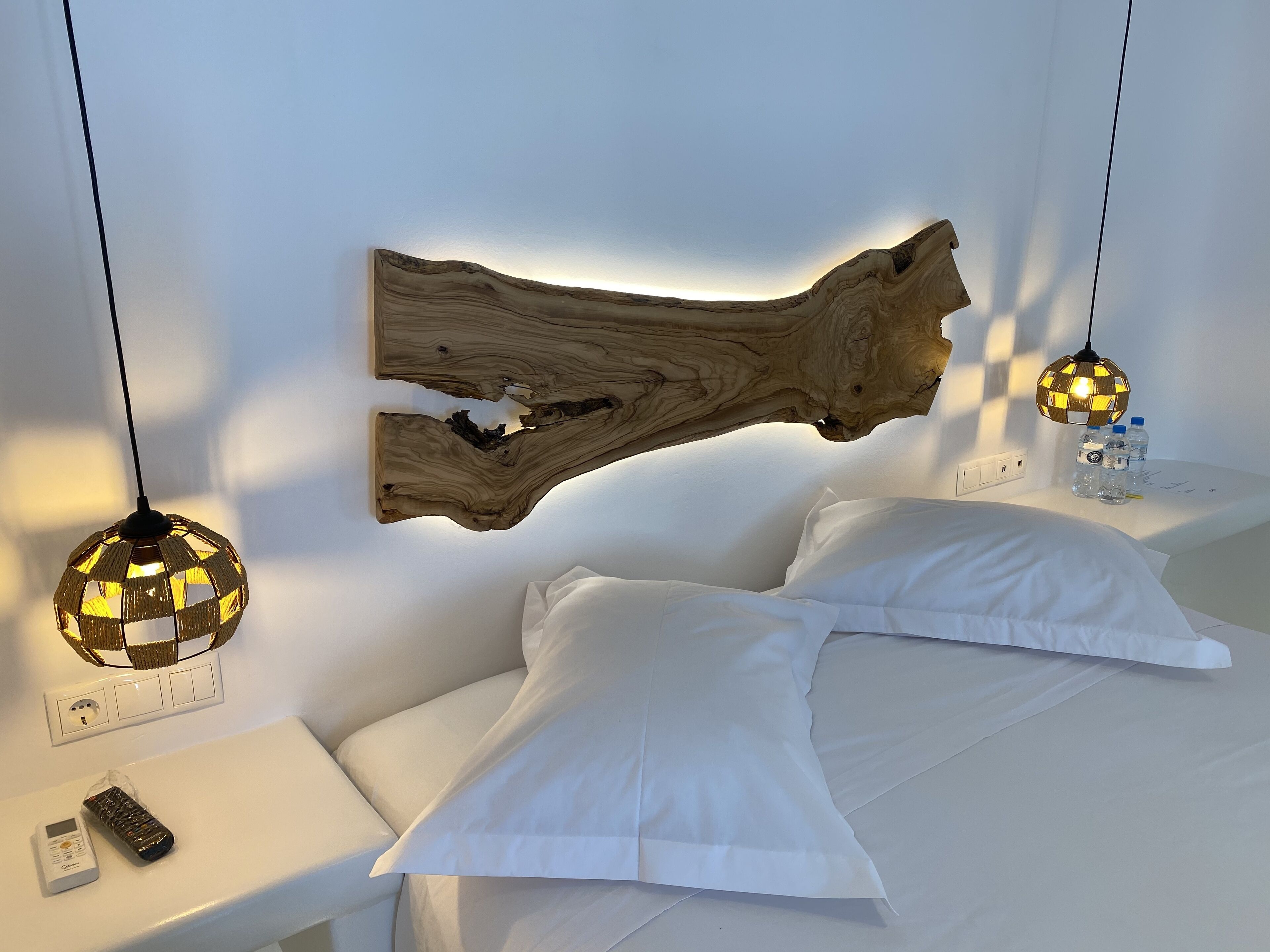 A modern bedroom with a large wooden wall art piece, two spherical lamps, a white bed, and a nightstand with a remote control and a water bottle.