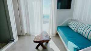 Une pièce avec un banc en bois, un canapé orné de coussins à rayures, des rideaux blancs et un téléviseur fixé au mur.