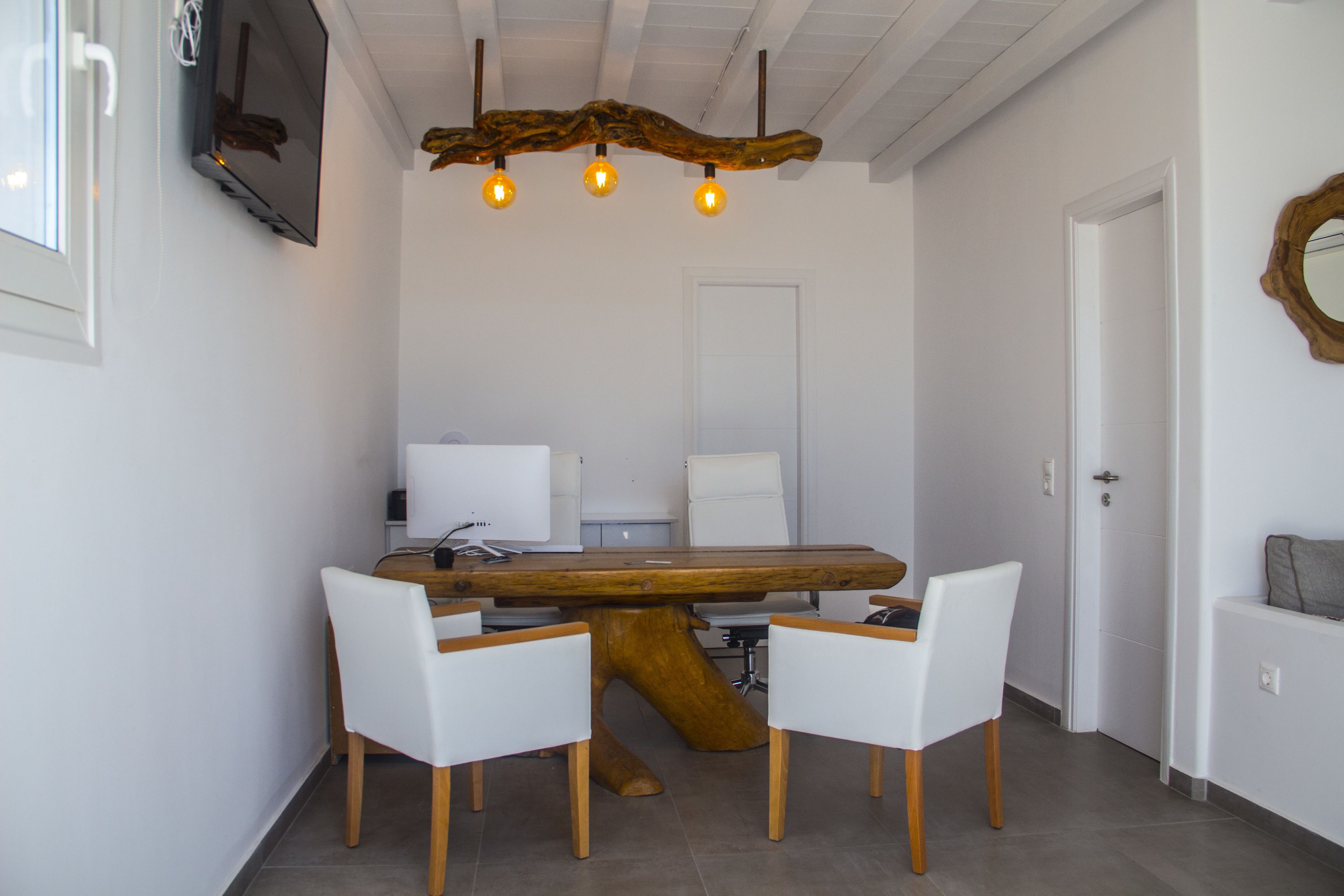 Un comedor con una mesa de madera, cuatro sillas blancas, un televisor montado en la pared y una lámpara en el techo.