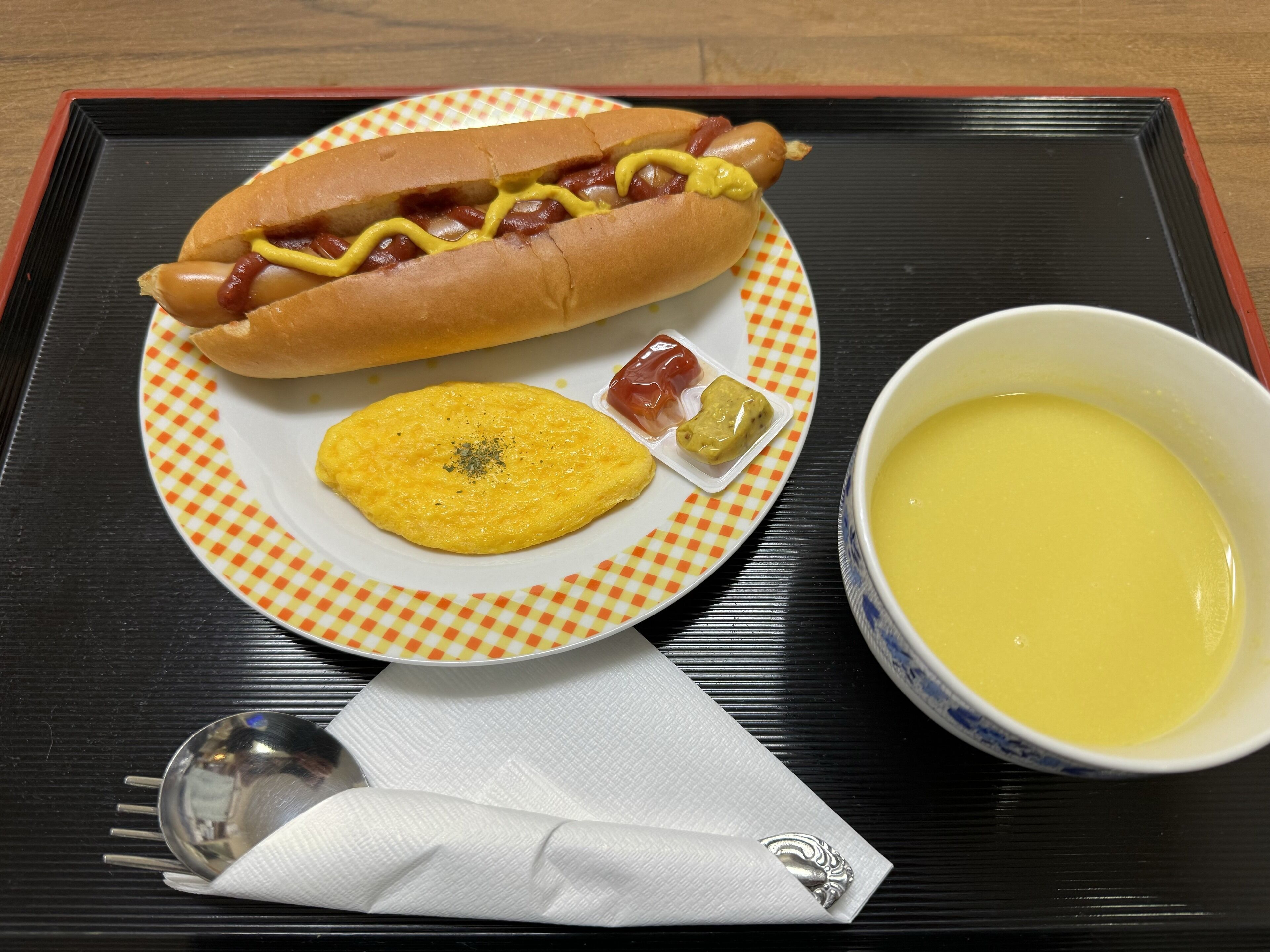 A plate with a hot dog, a small omelet, ketchup, mustard, and a bowl of soup.