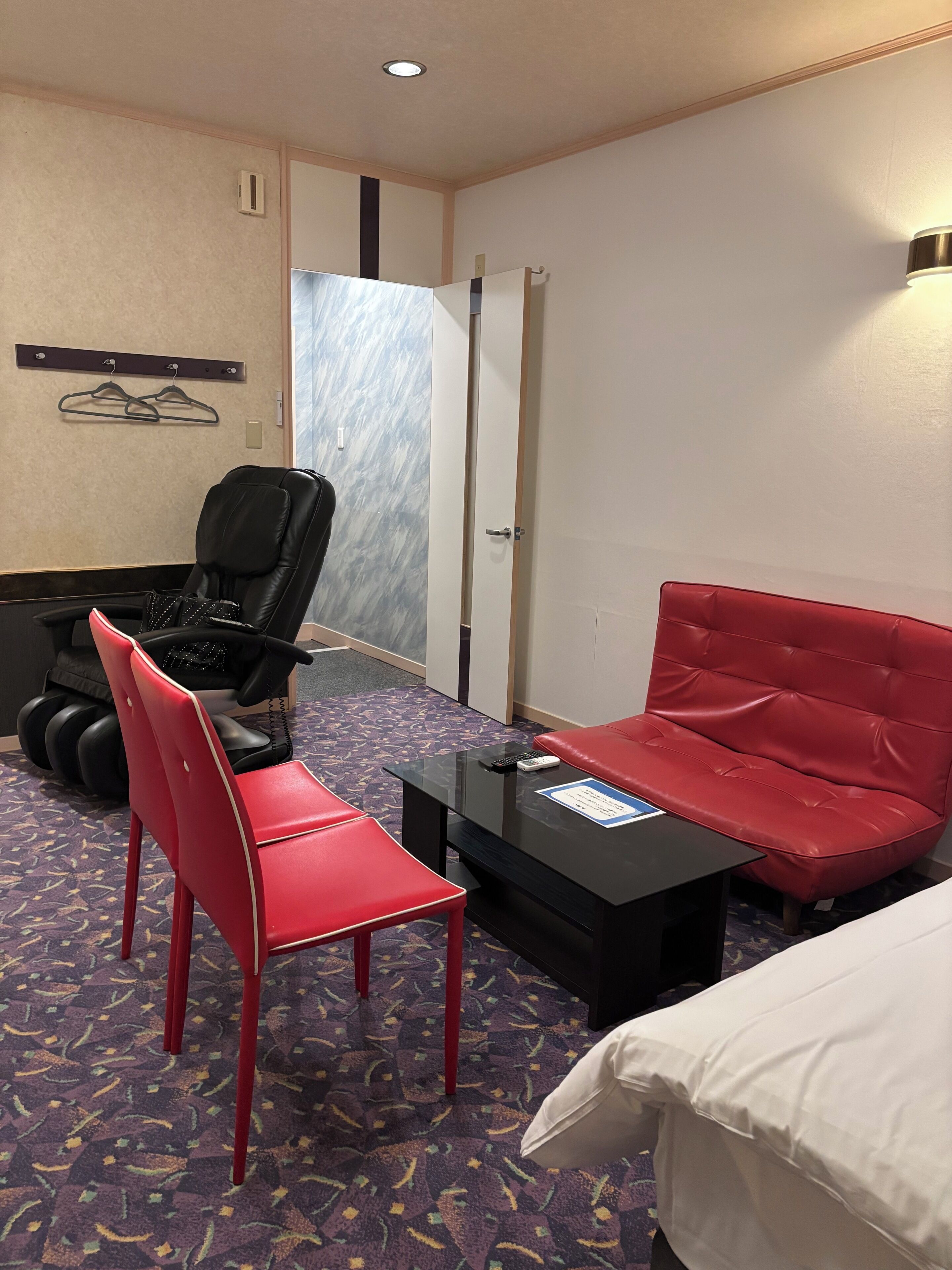 Habitación de hotel con una silla roja, una silla negra, una mesita, una cama y una puerta.