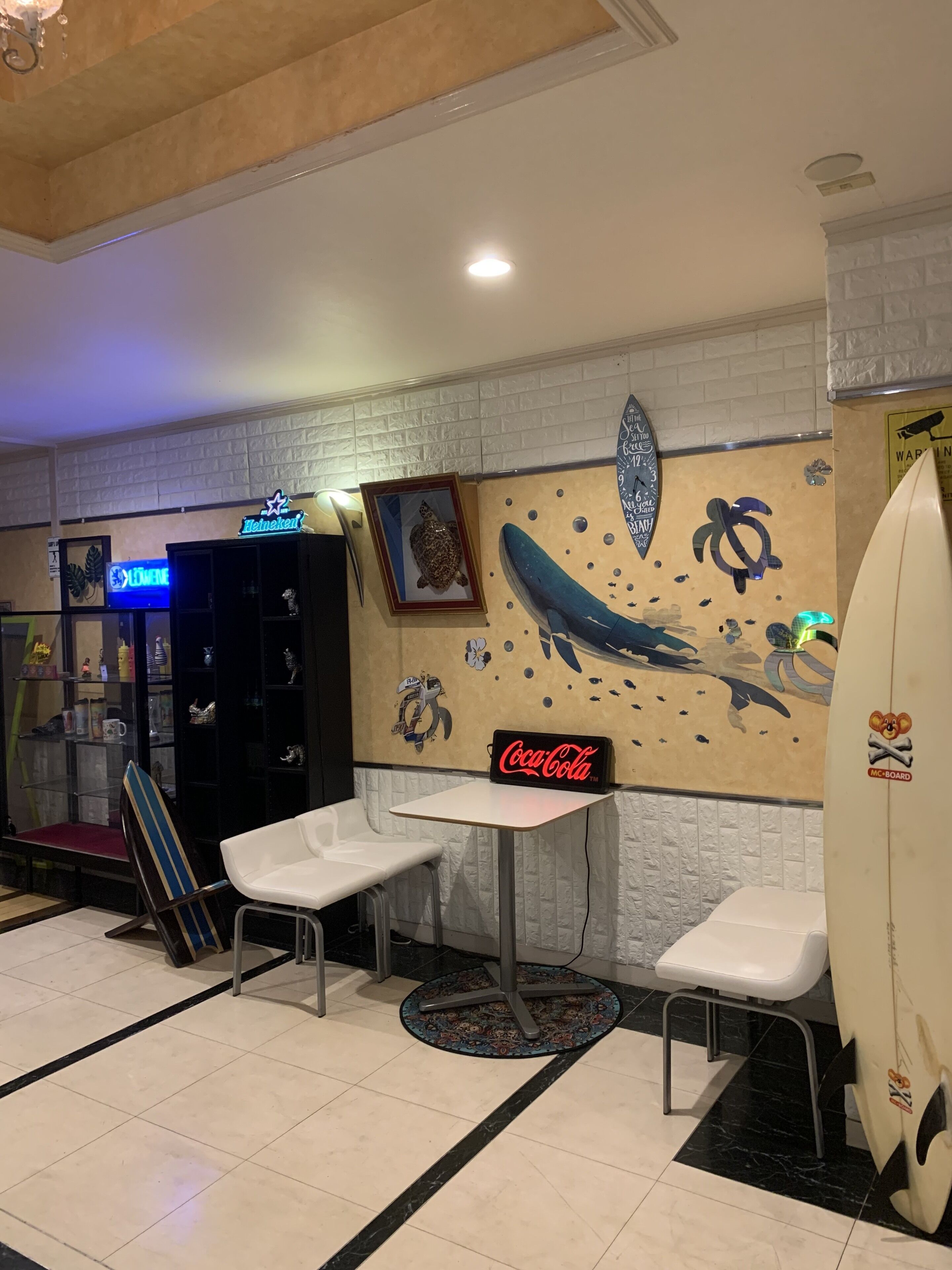 A room with a white brick wall, a framed turtle picture, a Coca-Cola sign, and a surfboard with a skull sticker.