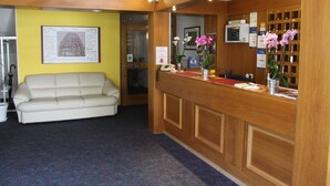 A reception area with a wooden desk, a white sofa, and a framed map on the wall. - Hotel Vitalesca (Neuschoenau)