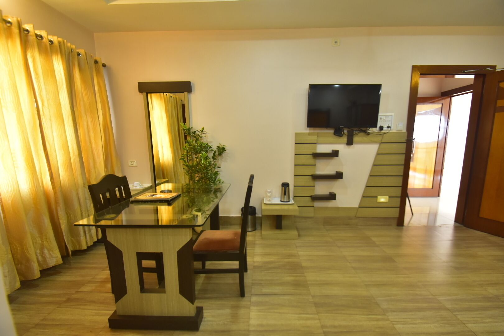 A modern living room with a glass dining table, wooden chairs, a sofa, a TV mounted on the wall, and a staircase leading to another room.