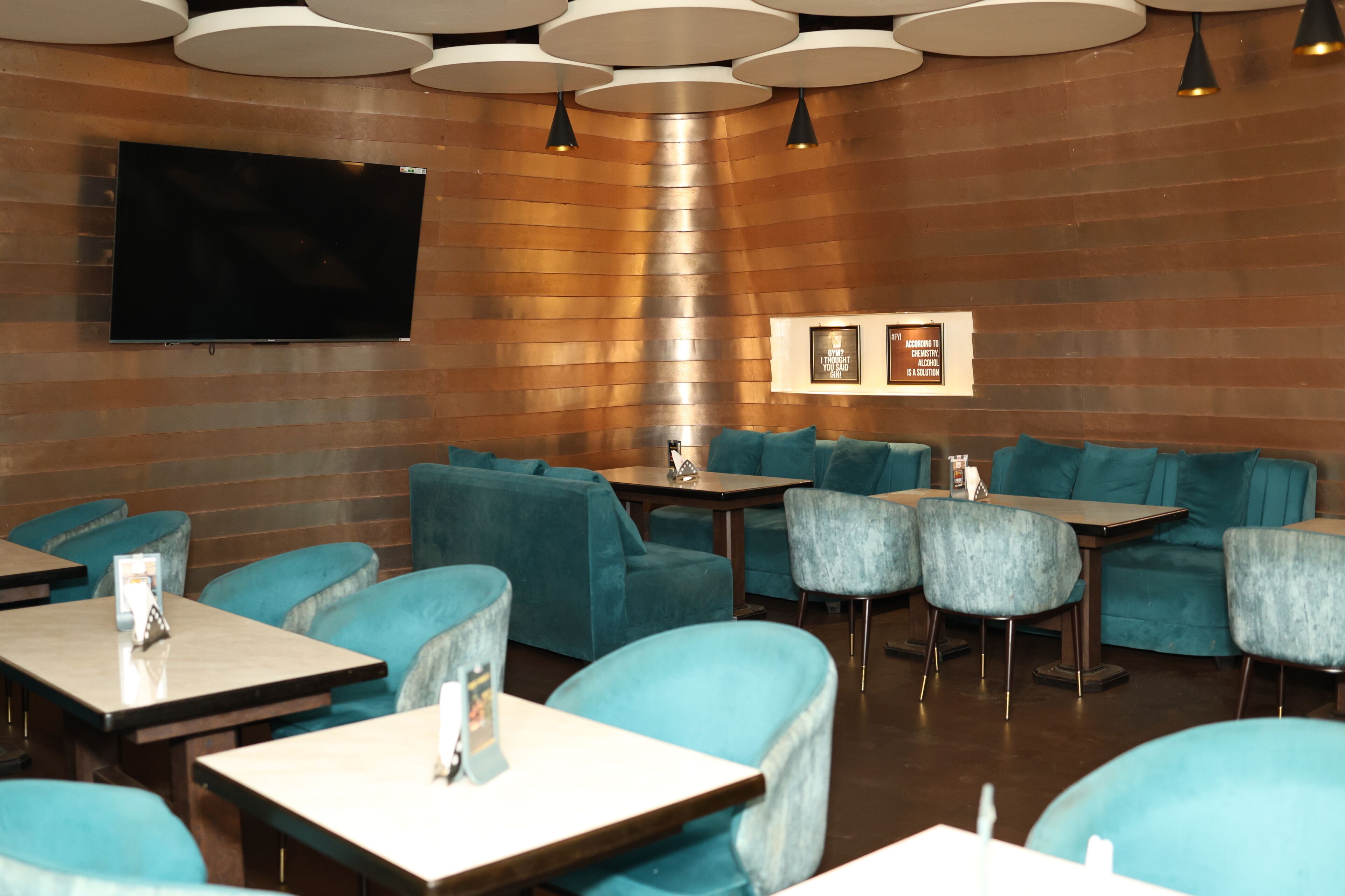 Un restaurante con asientos tapizados en color verde azulado, un televisor de pantalla plana montado en la pared e iluminación colgante.