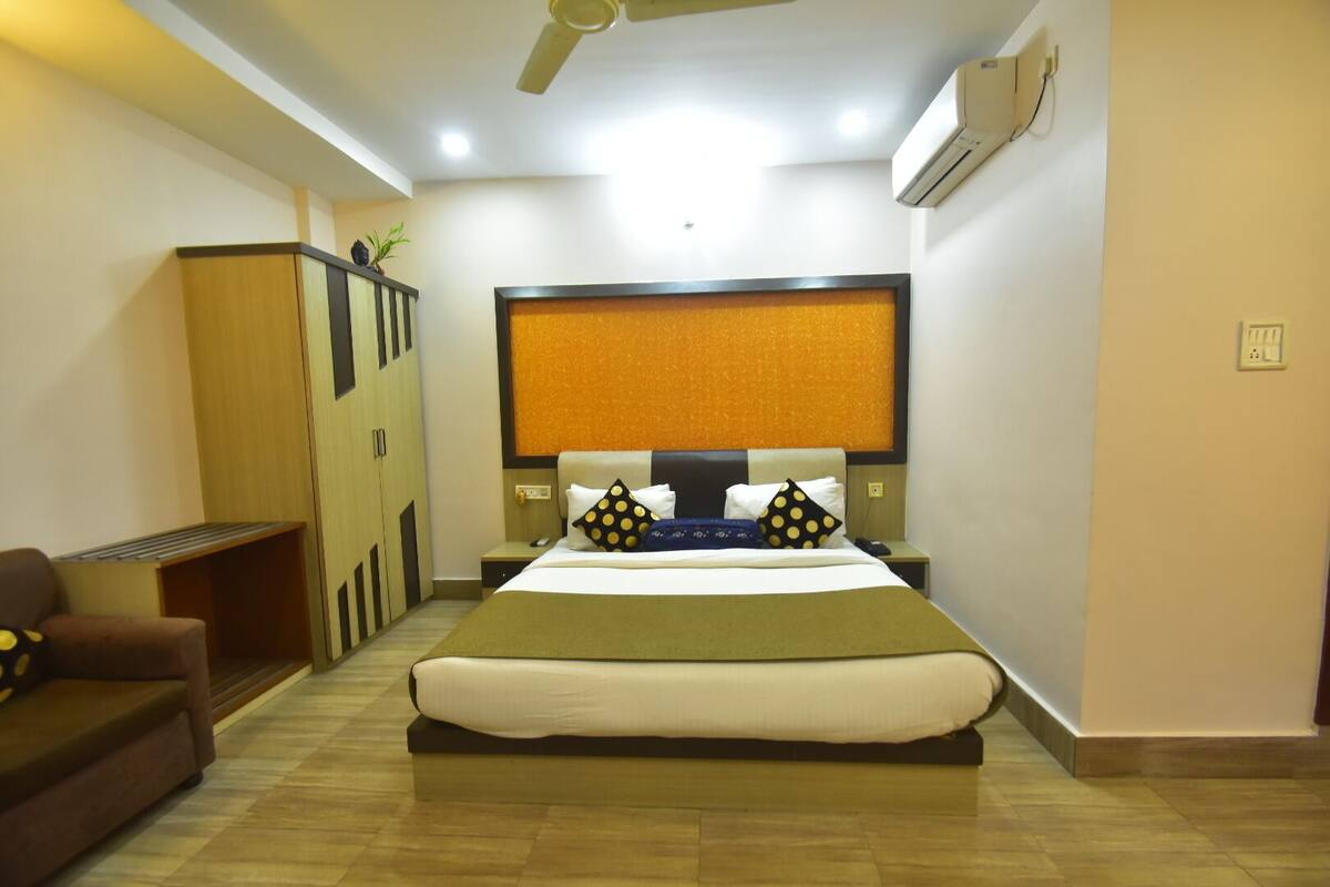 Quarto de hotel com uma cama, uma cadeira, uma mesa de cabeceira, ar condicionado e ventoinha no teto.