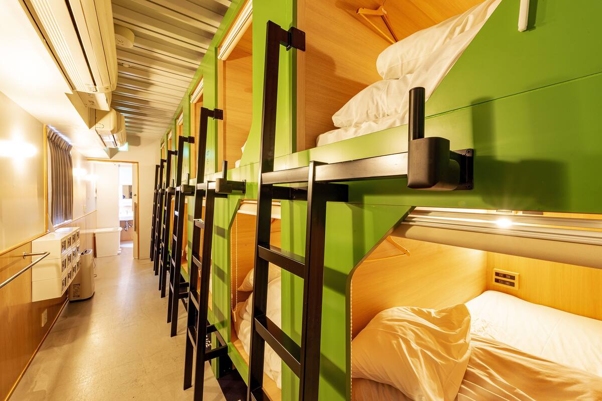 Varias literas con estructura verde y ropa de cama blanca, un rincón de cocina y un pasillo que conduce a una puerta.