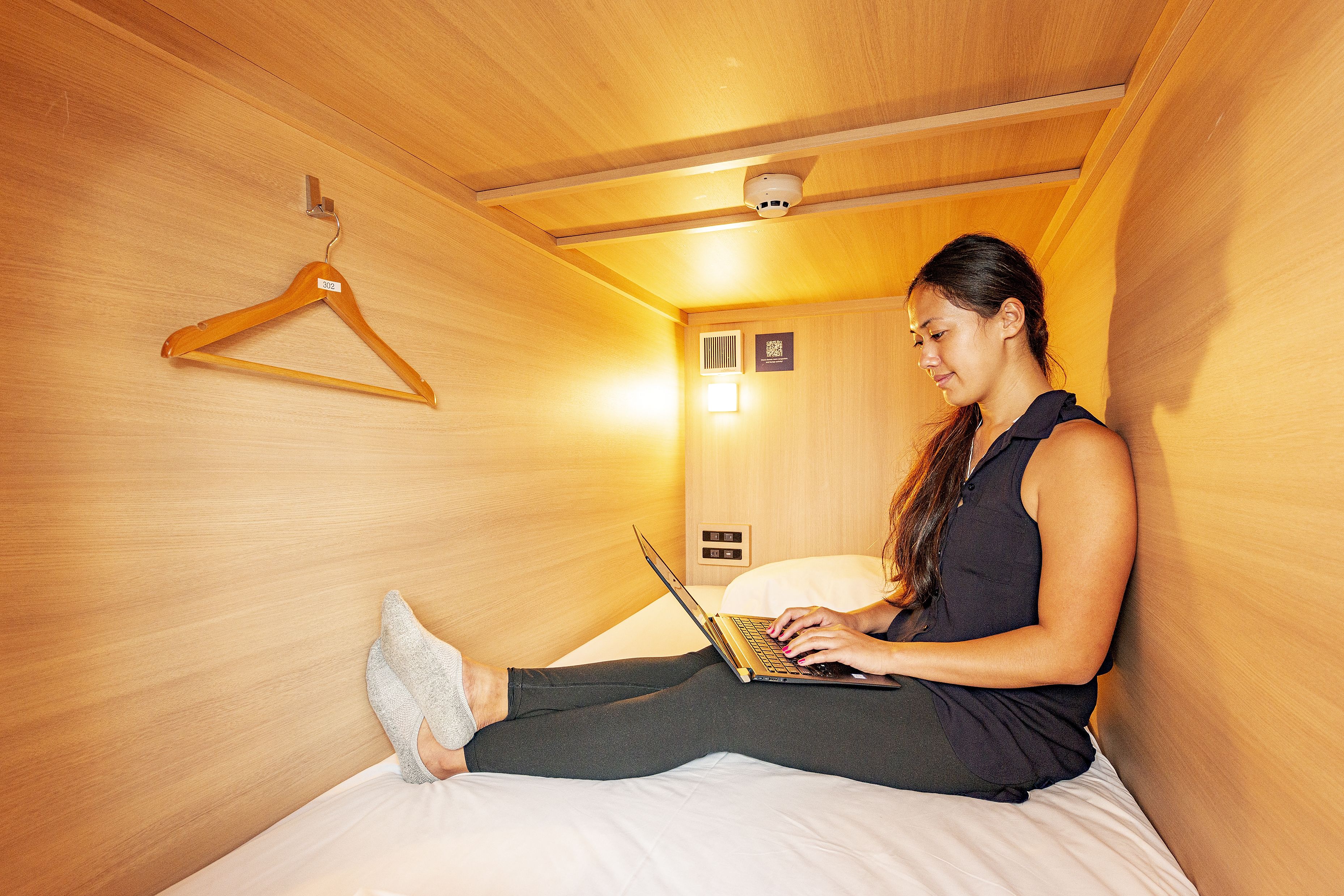 Een vrouw zit op een bed in een kamer met houten wanden en gebruikt een laptop.
