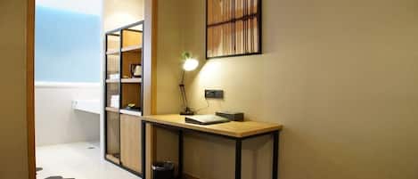 A hotel room with a wooden desk, a small trash bin, a wall-mounted lamp, and a framed picture.