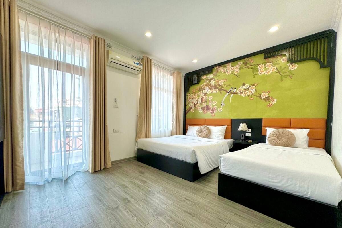 Une chambre d’hôtel avec deux lits, une grande fenêtre avec des rideaux, un appareil de climatisation et une fresque florale sur le mur.