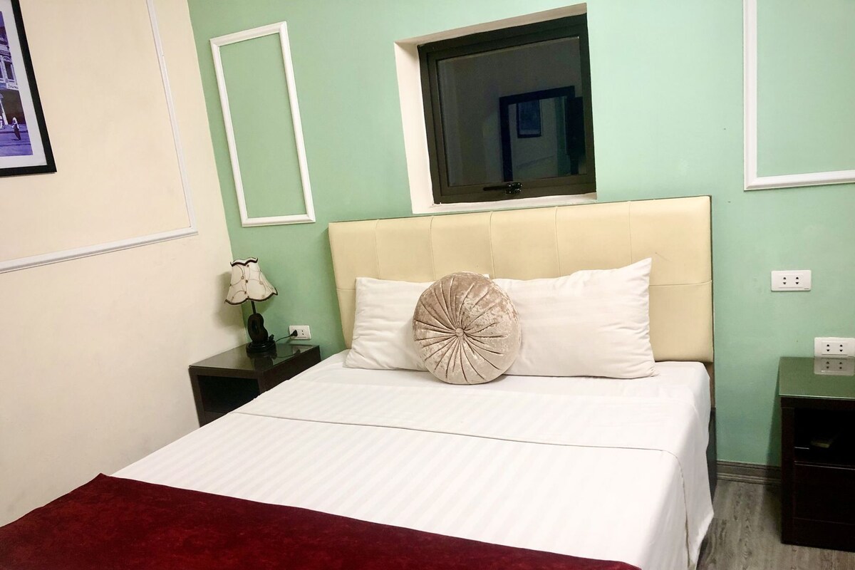 A neatly made bed with white bedding, a red throw blanket, a bedside lamp, and a nightstand with a small table.
