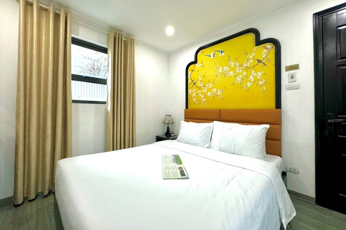 Une chambre d’hôtel avec un grand lit, des rideaux beiges, un mur jaune orné de motifs floraux et une porte noire.