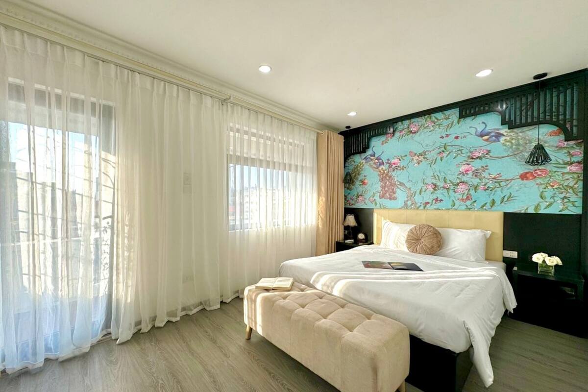 Une chambre avec deux lits, une grande fenêtre avec des rideaux légers, un plancher en bois et une fresque florale sur le mur.