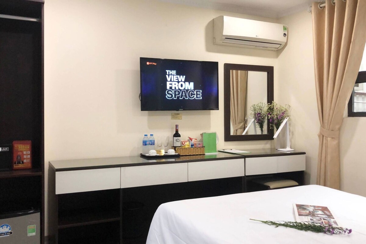 A hotel room with a bed, a desk with a flat-screen TV, a mirror, an air conditioner, and a window with curtains.