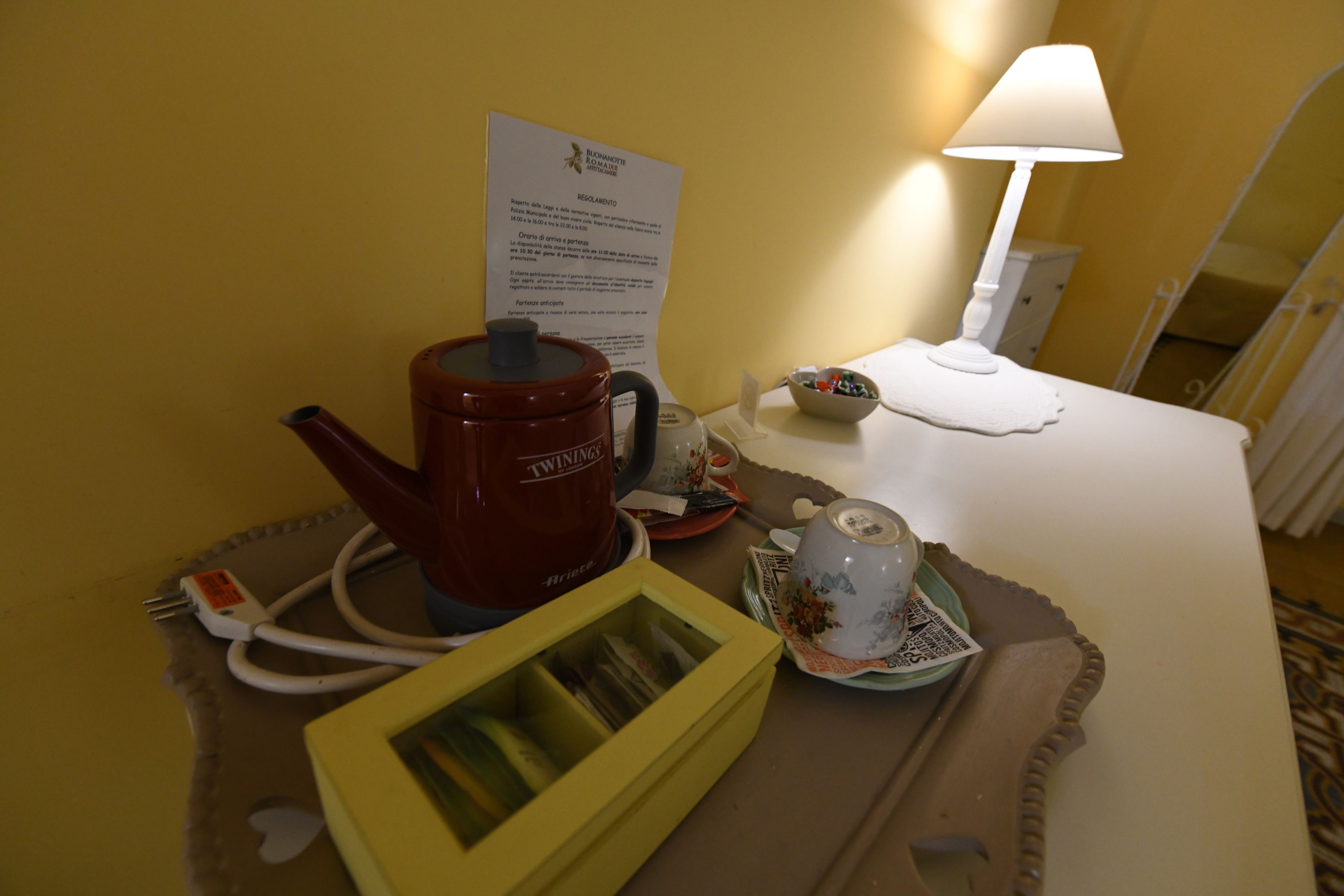 Habitación de hotel con hervidor rojo, una mesita con tetera, un bol de aperitivos, un cartel y una lámpara.