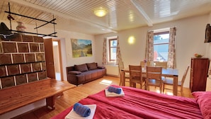 A cozy room with a wooden bench, a sofa, a dining table with chairs, and a bed with red bedding. - Jacob´s Resort House Podkoren (Kranjska Gora)