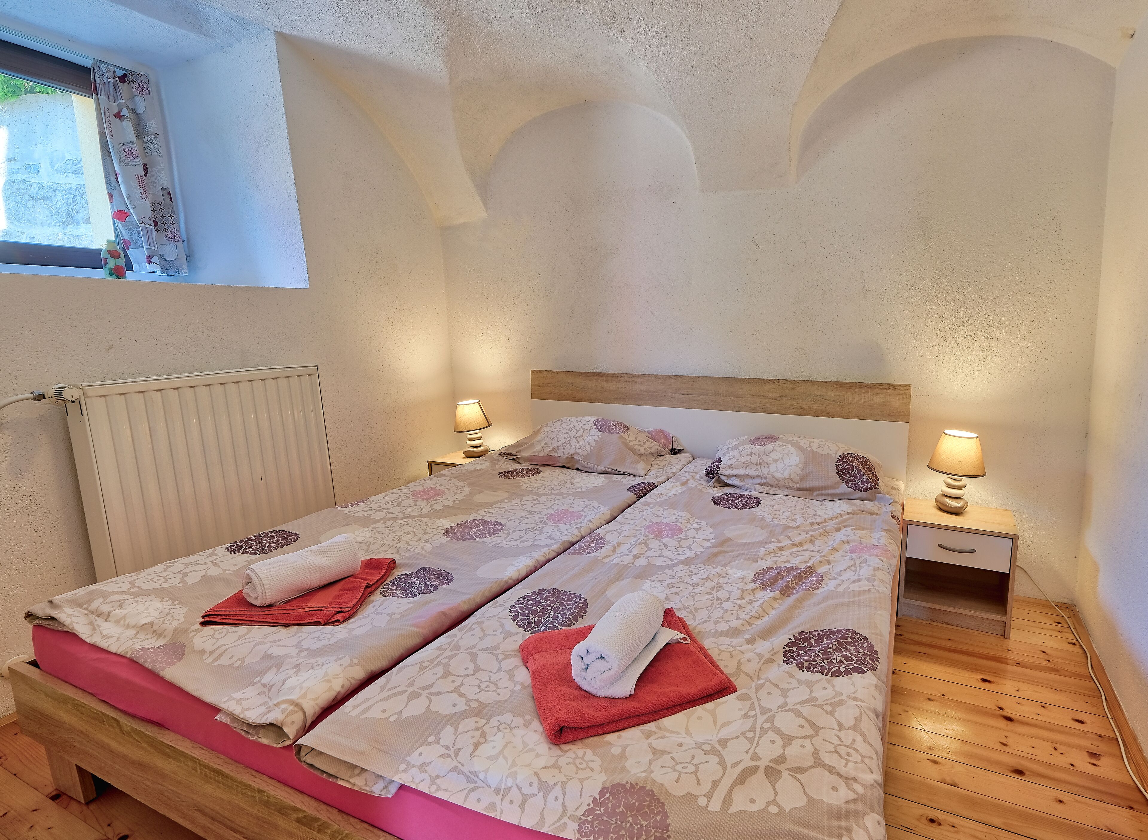 A double bed with floral bedding, two bedside tables with lamps, a wooden bench, and a window with curtains.