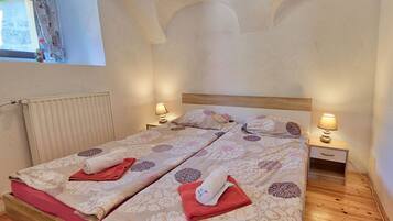 A double bed with floral bedding, two bedside tables with lamps, a wooden bench, and a window with curtains.