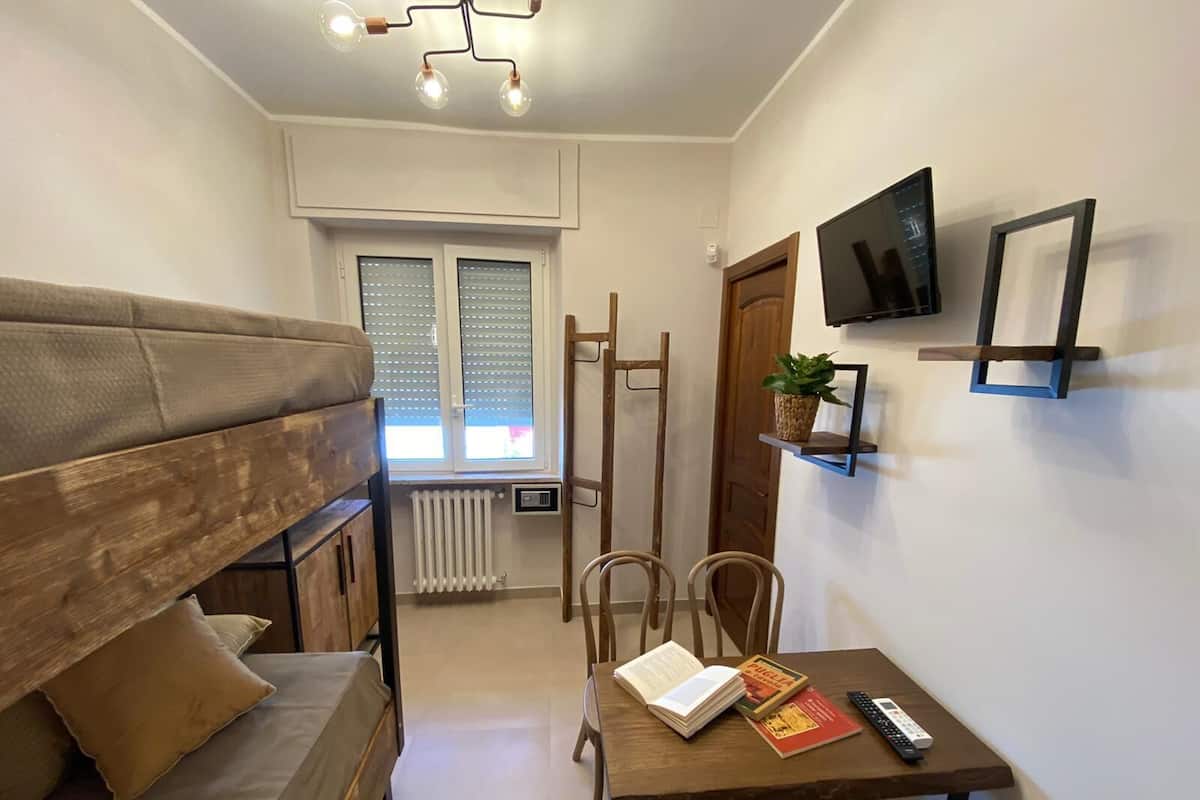 A cozy room with a wooden bunk bed, a table with books and a remote, a TV on the wall, and a window with blinds.