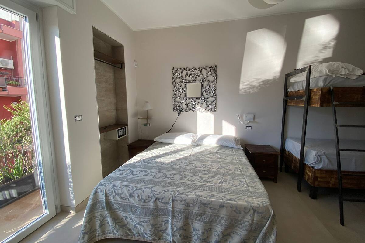 A bedroom with a bed, a bedside table, a lamp, a bunk bed, and a window with a view of a red building.