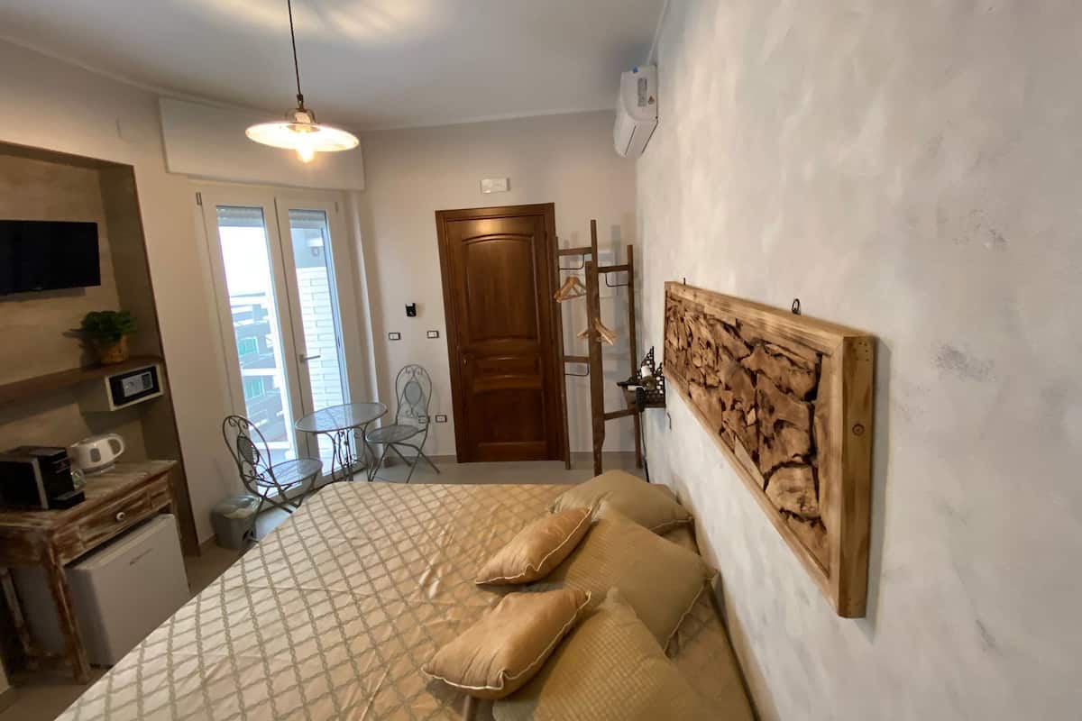A bedroom with a bed, a wooden headboard, a wall-mounted artwork, a small table with chairs, a desk with a computer, and a window with blinds.