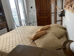 A neatly made bed with patterned bedding and two pillows, a small table with a chair, a wooden door, and a wall-mounted light fixture. - La Fontanella (Lecce)