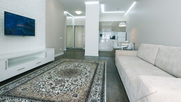 A modern living room with a white sofa, a flat-screen TV mounted on a white brick wall, a patterned rug, and a kitchen area in the background. - VIP Apartment Company (Kyiv)