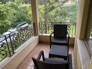 A balcony with a wicker chair and table, a metal railing, and a view of greenery and a parked car.